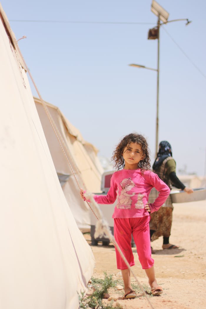 hero-img-02 A young girl in bright clothing stands next to a tent in a Syrian refugee camp under a clear sky.